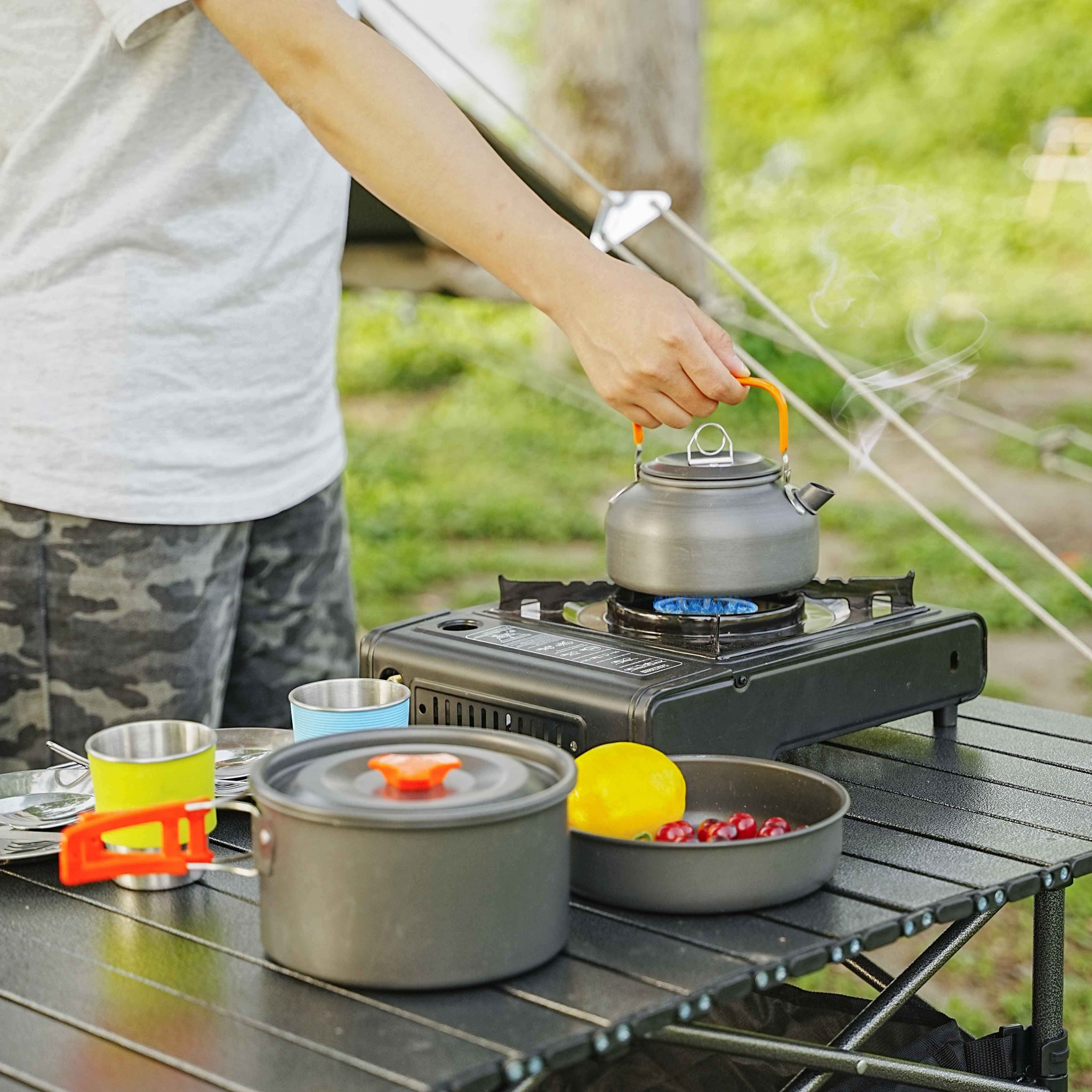 Person cooking outdoors using a portable stove with pots and a kettle.