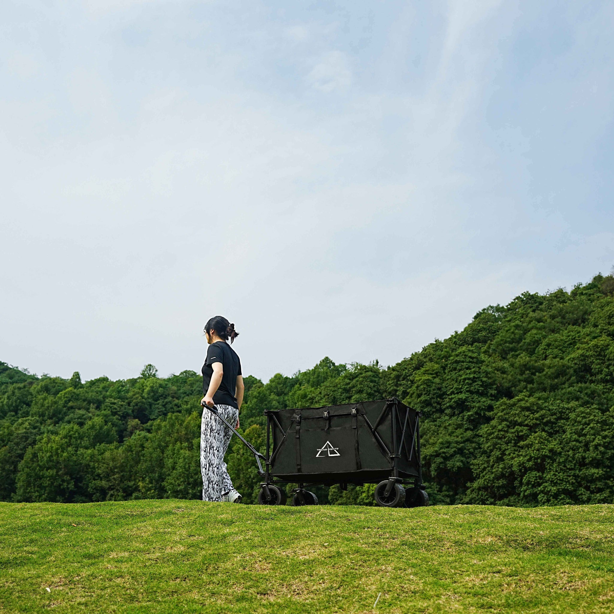 Person pulling a black cart with a logo on a grassy field with trees in the background