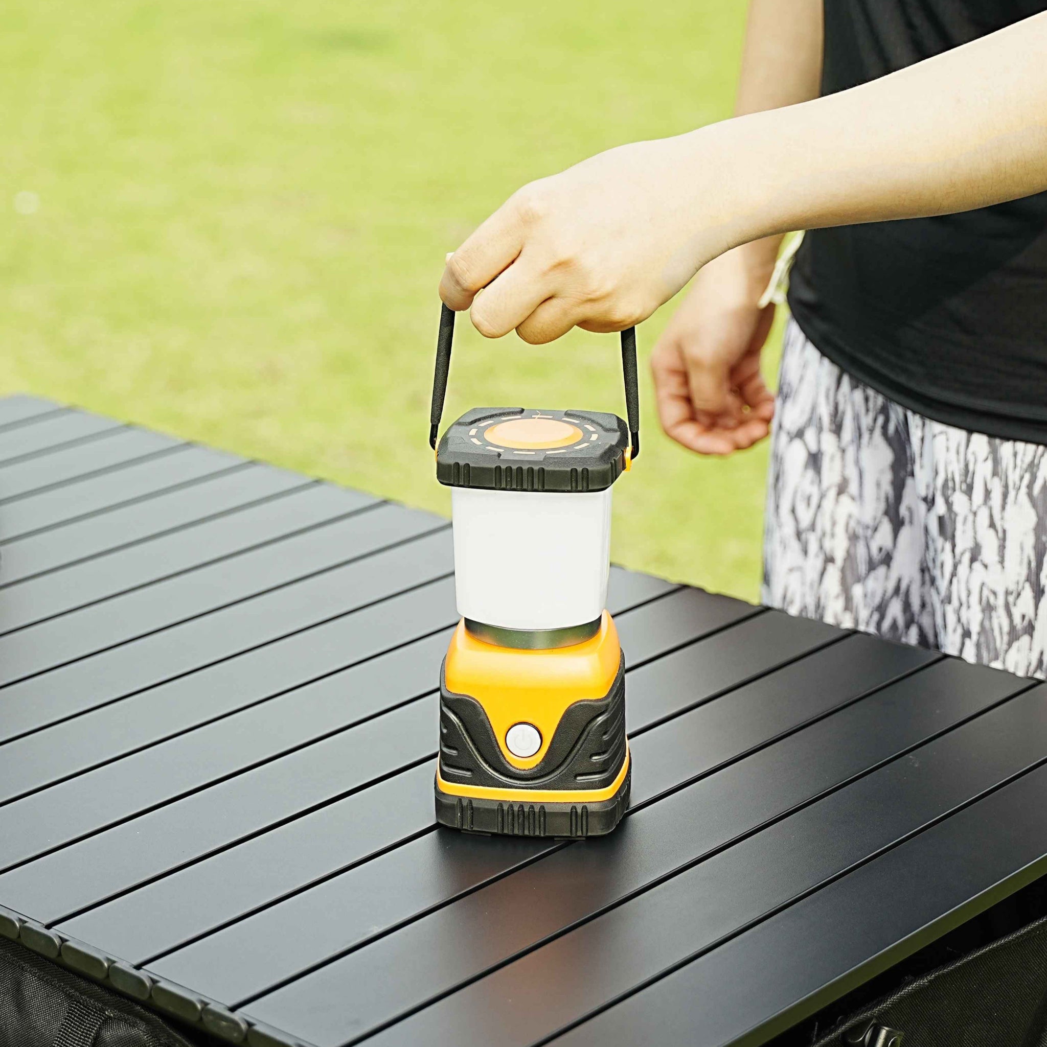 Hand holding a yellow and black camping lantern on a wooden surface with a blurred green background