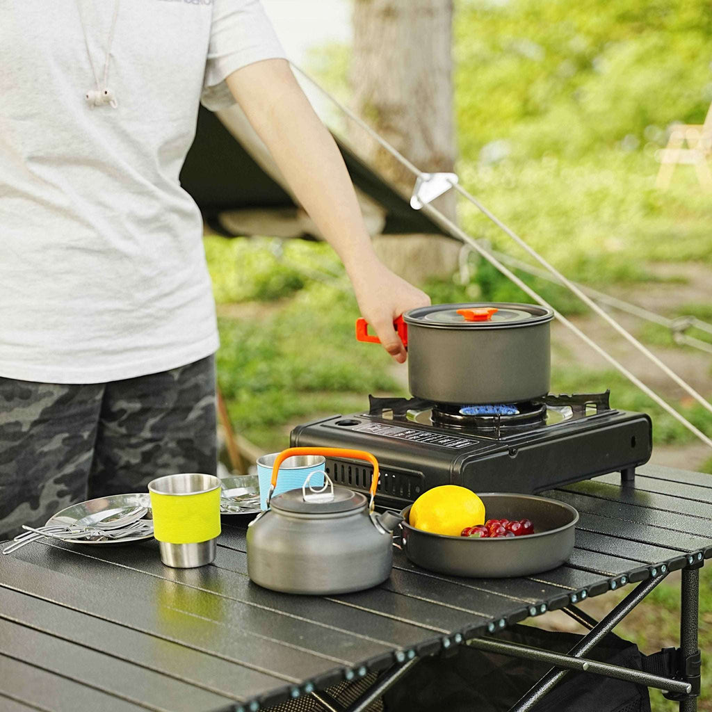 Person cooking outdoors with a portable stove and cookware on a camping table.