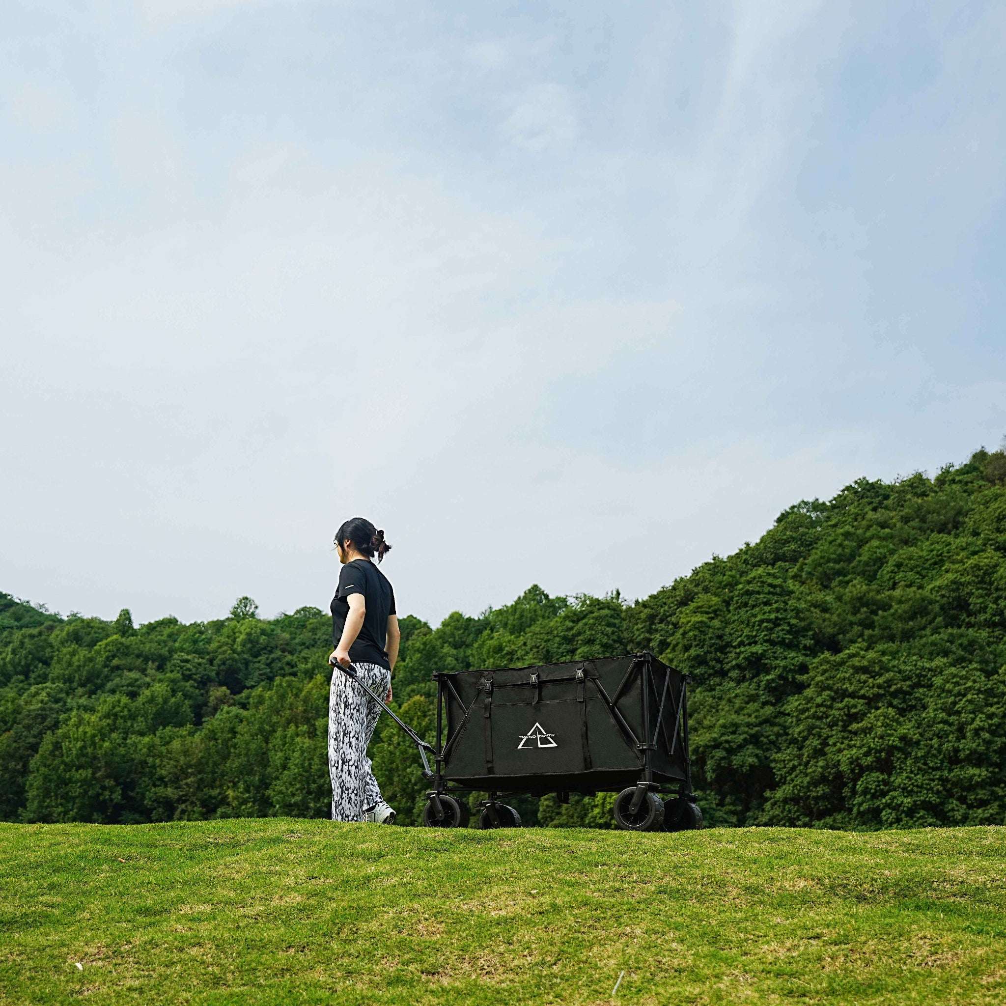 Person pulling a black cart with a logo on a grassy field with trees in the background